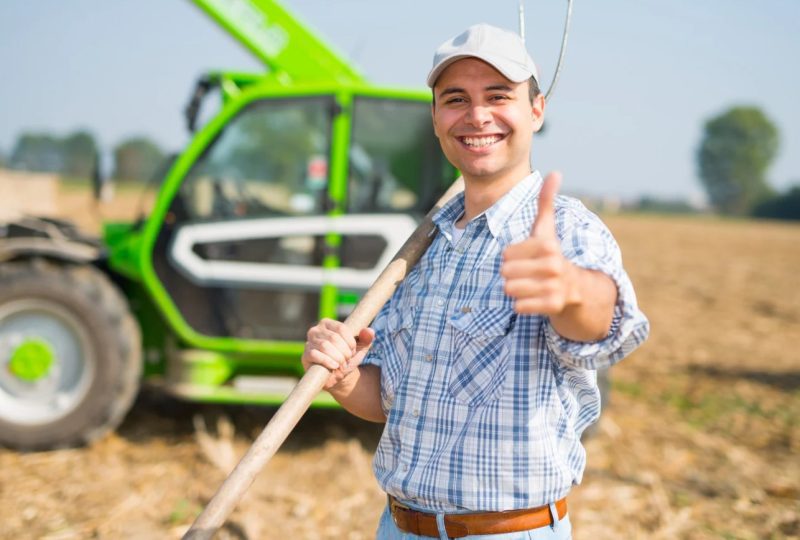 Młody rolnik z widłami na tle zielonego ciągnika.