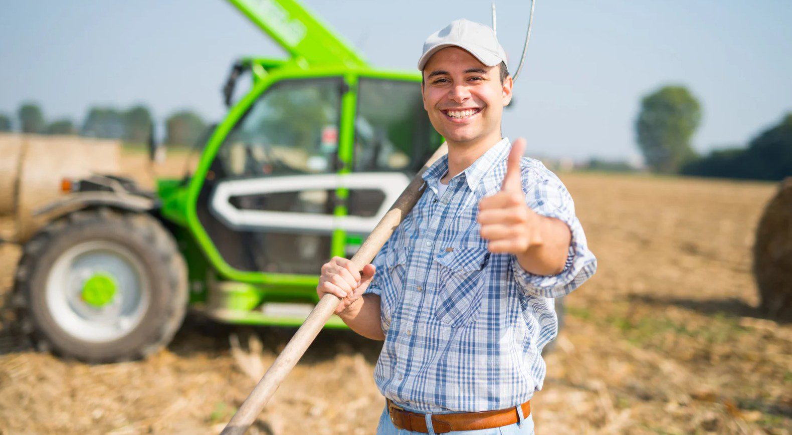 Młody rolnik z widłami na tle zielonego ciągnika.